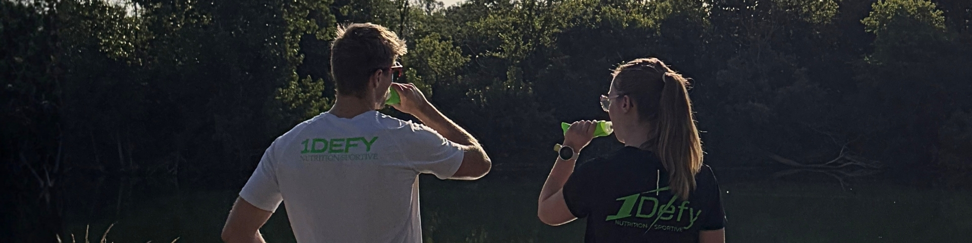 Photographie d'un homme et d'une femme de dos, avec un t-shirt 1DEFY Nutrition sportive et buvant de l'eau, 2 sportifs au bords de l'eau, photographie utilisée pour l'article de blog sur les boissons isotoniques de 1Defy Nutrition Sportive Made in Bourgogne
