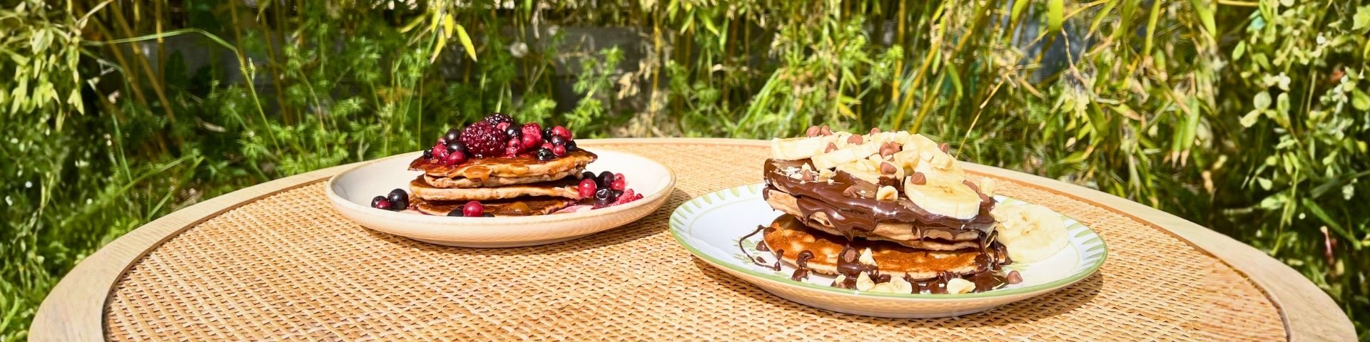 Pancakes protéinés posés sur une table, un pancake avec des fruits rouges en topping et un pancakes avec des bananes, du chocolat et des noisettes, photo pour la recette : Pancakes protéinés